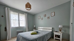 Ein Schlafzimmer mit einem Bett, einem Nachttisch, einer Lampe, einem Fenster mit Vorhängen und Bildern an der Wand.