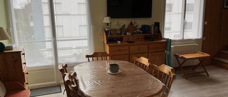 A dining area with a wooden table and chairs, a TV mounted on the wall, and a window with a view of the outside.