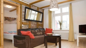 A cozy living room with a brown leather sofa, a wooden coffee table, a flat-screen TV mounted on a brick wall, and a window with curtains. - Apartment Westwind with view of churches - Gohr- apartments (Stralsund)