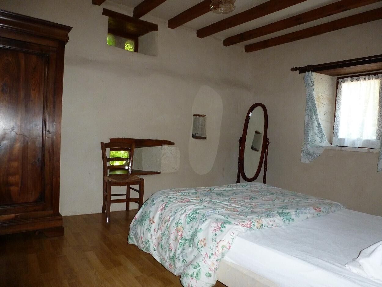 Ein Schlafzimmer mit einem hölzernen Bett, einem Sessel, einem Holzschrank, einem Fenster mit Vorhängen und einem kleinen Oberlicht.