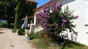 Un edificio blanco con techo de tejas, camino de grava y un arbusto morado en flor.
