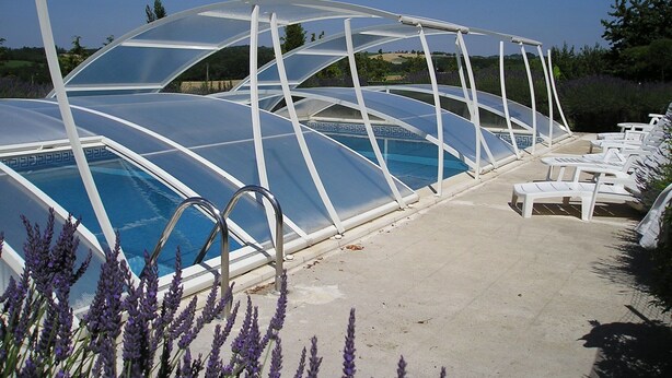 Un área de piscina con una estructura de vidrio, sillas reclinables blancas y plantas de lavanda.