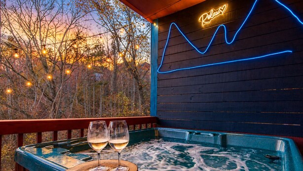 A hot tub with two glasses of wine, a neon sign reading 'Relax,' and a view of trees at sunset. - Smoky Mtn Cabin -Theater, Hot Tub, Games-9 min to Dollywood (Sevierville)