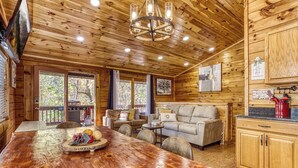 A wooden cabin interior with a dining table, two sofas, a kitchen area, and a large window overlooking the outdoors. - Smoky Mtn Cabin -Theater, Hot Tub, Games-9 min to Dollywood (Sevierville)