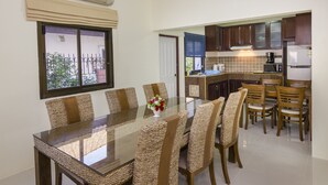 A dining area with a glass-top table and woven chairs, a kitchen with wooden cabinets, and a view through a window. - RATANA VILLA - PATTAYA HOLIDAY HOUSE - WALKING STREET (Muang Pattaya)