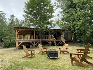Eine Holzhütte mit einer Terrasse, umgeben von Bäumen. Im Vordergrund stehen vier Adirondack-Stühle und ein schwarzer Grill.