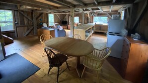 A rustic cabin with a wooden dining table, two chairs, a kitchen area with a stove, and a large window providing natural light. - Secluded trailside cabin for 8 people on Kingdom Trails  (Burke)