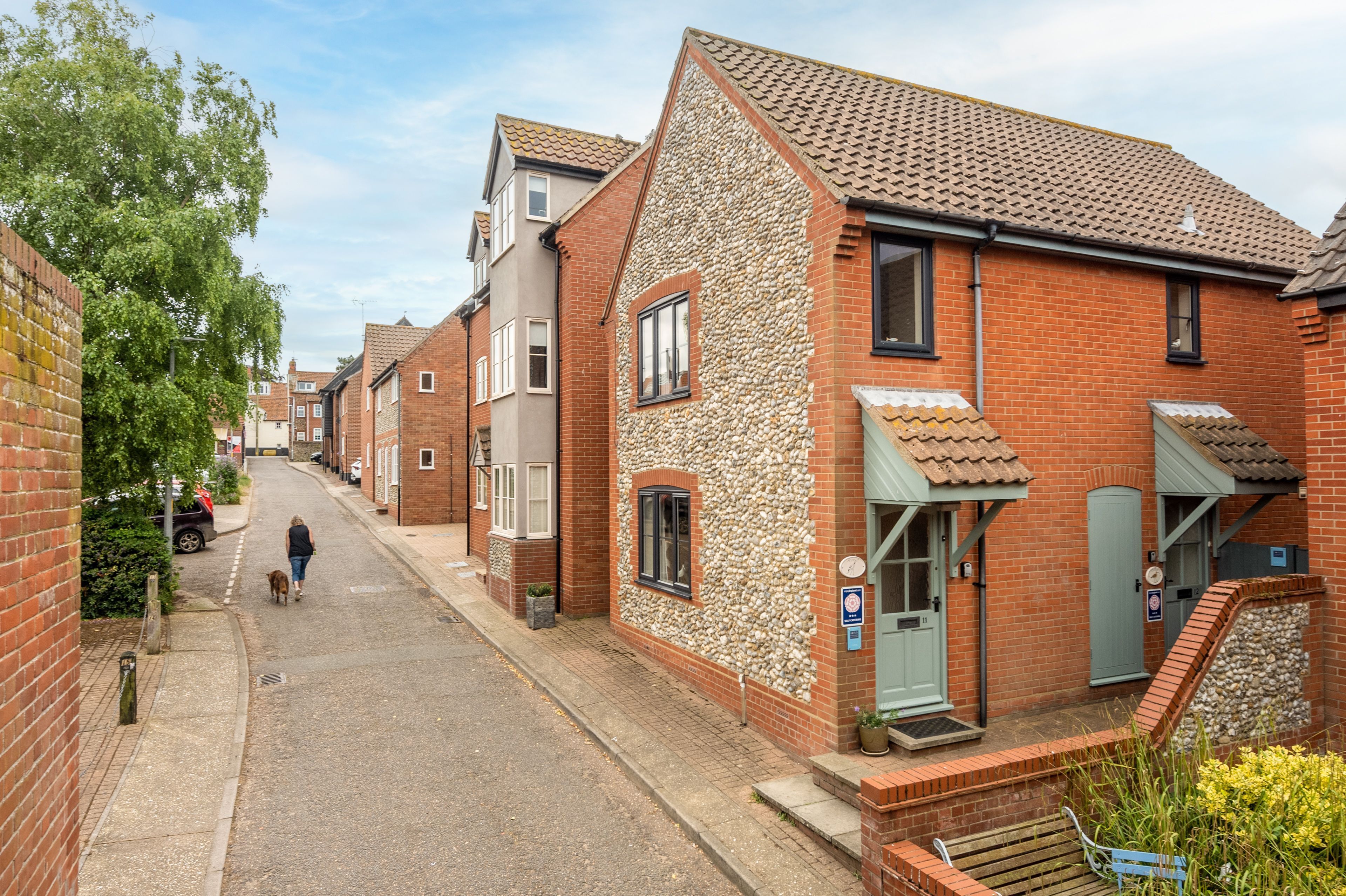 Une rue étroite bordée de maisons à étages, avec des façades en briques et en pierre, une personne promenant un chien et un ciel dégagé.
