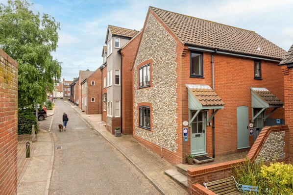 A narrow street lined with terraced houses, featuring brick and stone facades, a person walking a dog, and a clear sky.