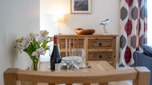 A dining table with a vase of flowers, a wine bottle, and glasses. There is a wooden cabinet with a bird sculpture and a framed picture on the wall behind. - Avocet Apartment, Wells-next-the-Sea, Norfolk (Wells-Next-The-Sea)