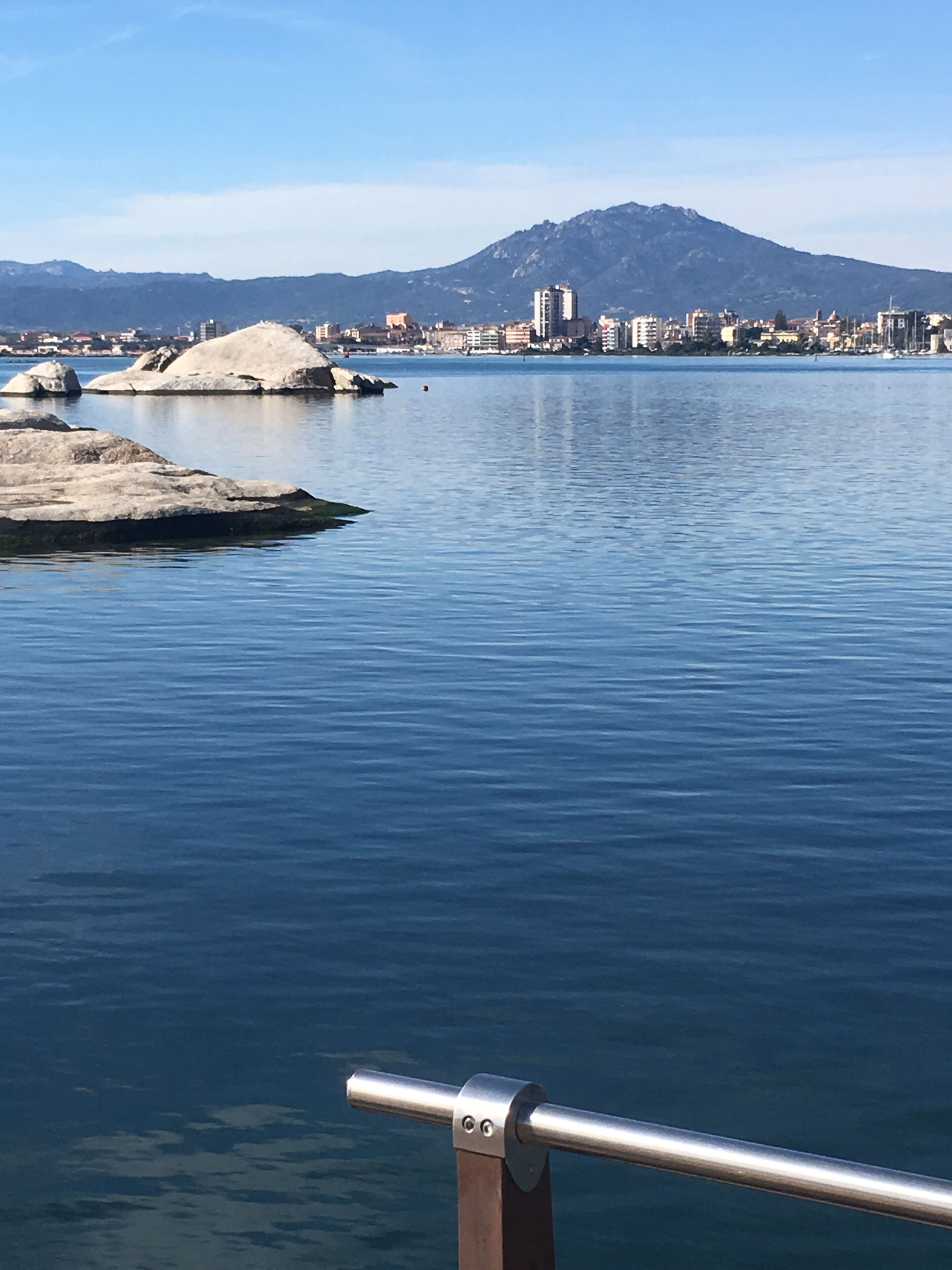A calm waterfront with large rocks, a metal railing, and a cityscape with mountains in the background.