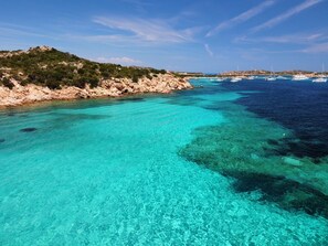 A coastal landscape with clear turquoise waters, rocky terrain, and boats in the distance.
