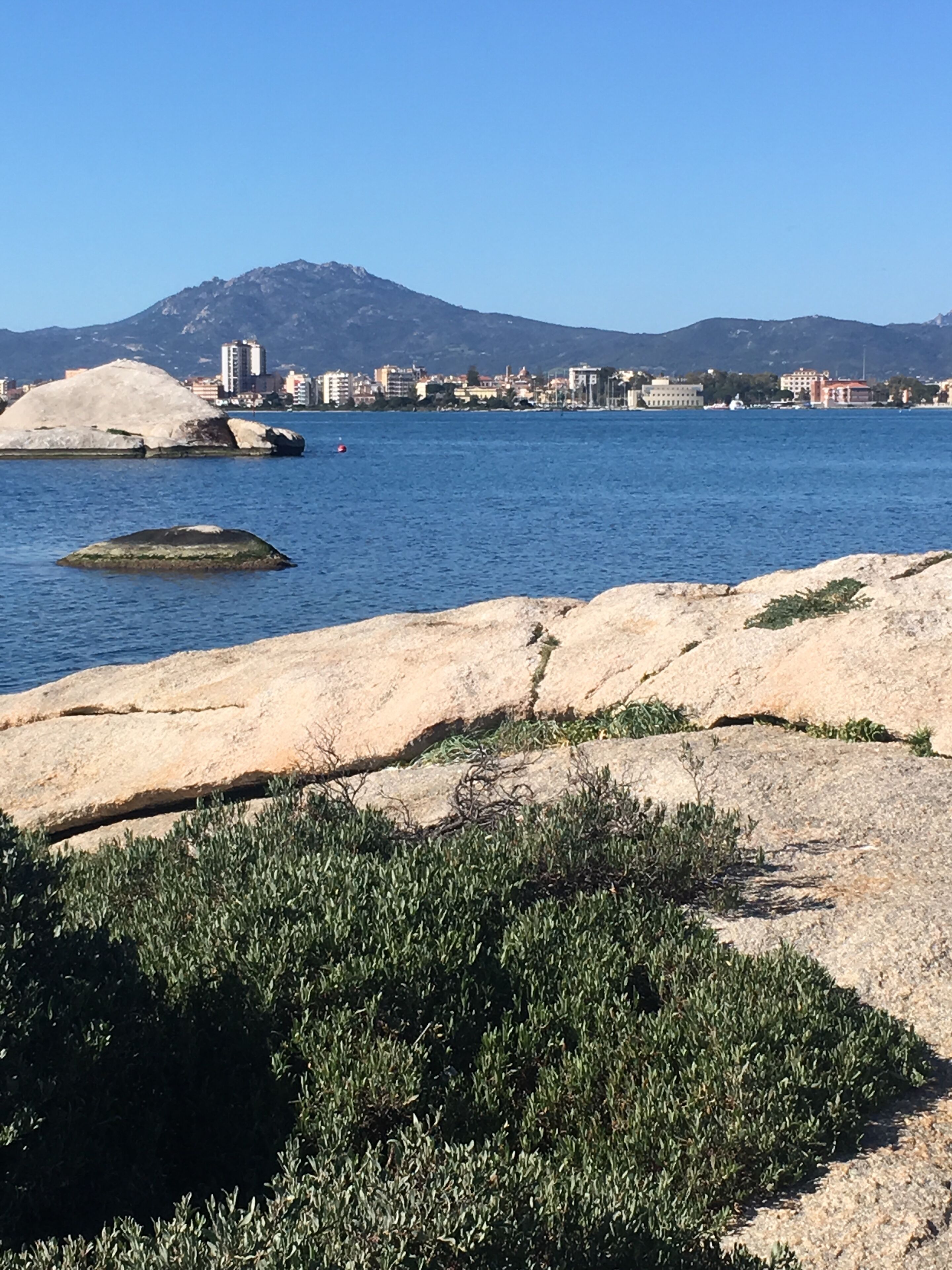 Paisatge costaner amb costes rocoses, mar blau clara i una ciutat llunyana amb edificis i muntanyes.
