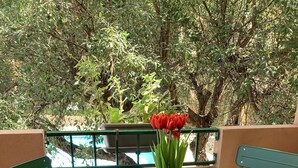 Un pequeño balcón exterior con una mesa verde y dos sillas verdes, cada una con un cojín. En la mesa hay una maceta blanca con flores rojas.