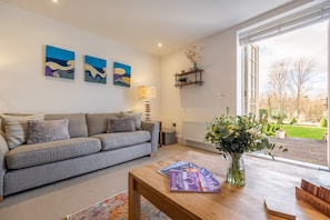 Un salon moderne avec un canapé gris, une table basse en bois sur laquelle sont posés des magazines, et un vase de fleurs. Deux tableaux sont accrochés au mur, et une étagère expose des objets décoratifs.