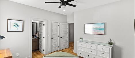 A bedroom with a bed, a ceiling fan, a dresser, a TV, and a door leading to a bathroom.