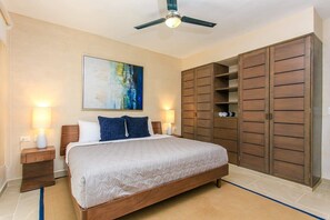 A bedroom with a bed, bedside lamps, a ceiling fan, a large wardrobe, and a painting on the wall.