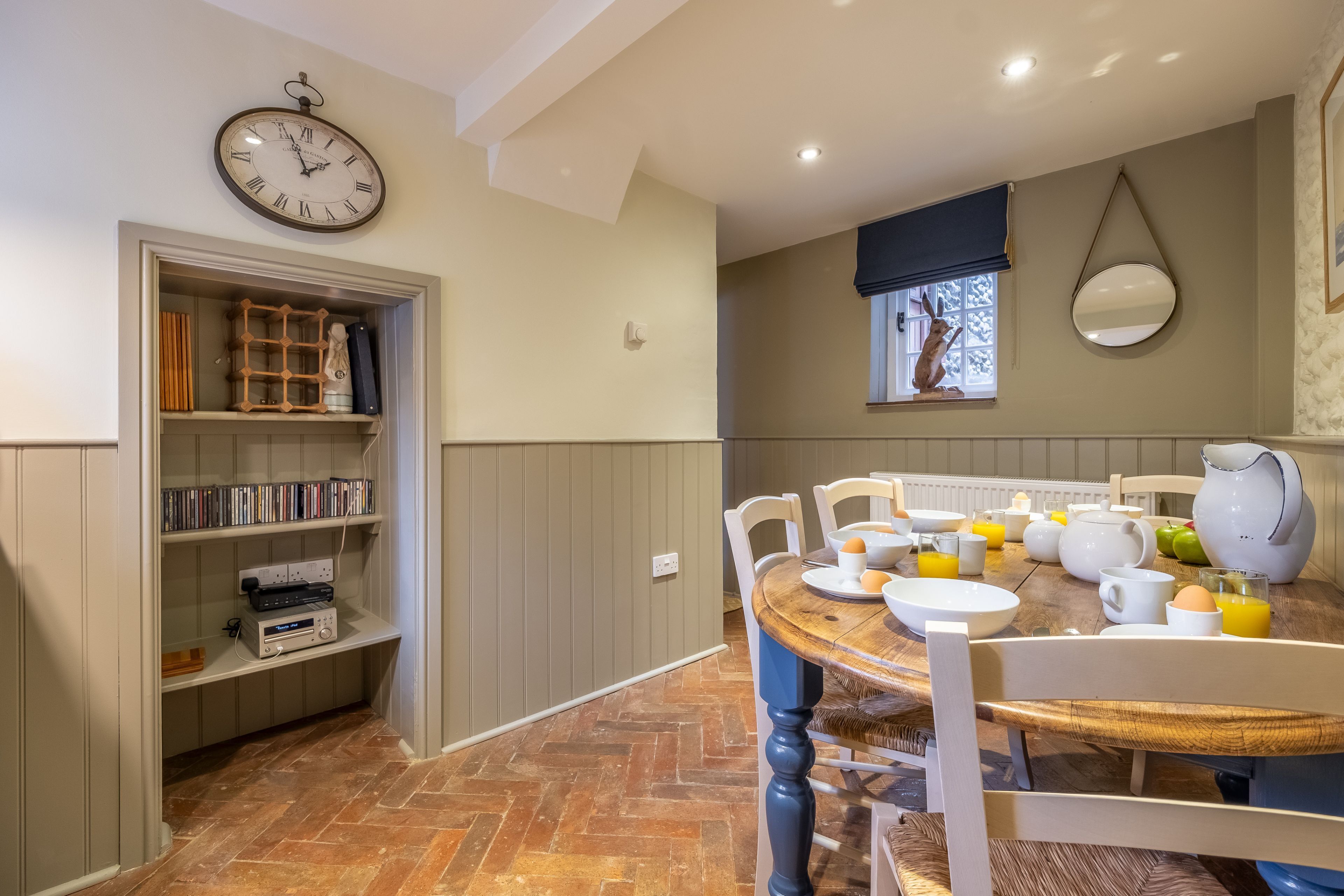 Uma sala de jantar com uma mesa de madeira, cadeiras brancas e um relógio na parede.