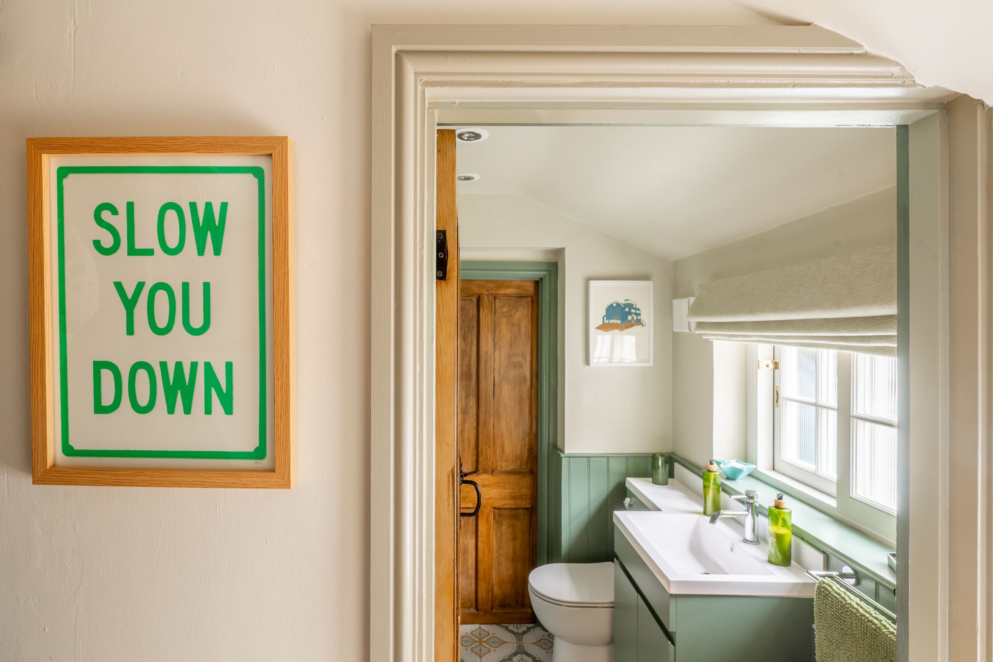 Un bagno con il water, il lavandino e un cartello incorniciato con su scritto 'SLOW YOU DOWN'.