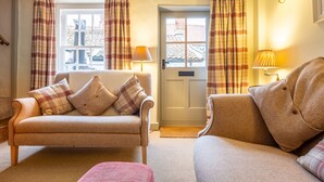 A cozy living room with a beige sofa, a matching armchair, and a small table. There are plaid curtains on the windows, and a lamp on the wall.