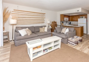 A living room with a grey sofa, a white coffee table with books, and a kitchen area with wooden cabinets and white appliances.