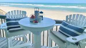 Une terrasse avec deux fauteuils Adirondack blancs, une petite table ronde et un coin café. La terrasse offre une vue sur une plage, avec l’océan en toile de fond.