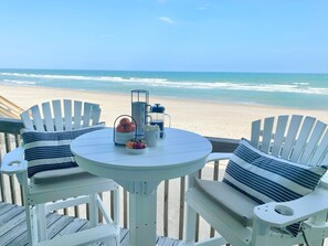 A deck with two white Adirondack chairs and a small round table with a coffee maker, mug, and fruit. The deck overlooks a beach with waves and a clear sky.
