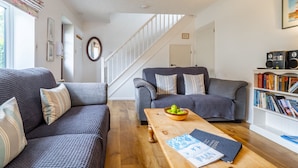 A living room with two grey sofas, a wooden coffee table with a bowl of fruit, a bookshelf, and a staircase.