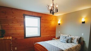 A bedroom with wooden walls, a large window, a bed with patterned bedding, a wooden dresser, and a chandelier. -  30 Mins to Waterton Gate! Relax & Enjoy the View of Chief Mountain & Sunsets (CARDSTON)