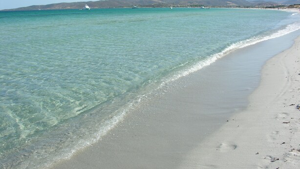 Ein Strand mit klarem türkisblauem Wasser, einem sandigen Ufer und Bergen im Hintergrund.