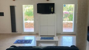 Ein geräumiges Zimmer mit einem großen Bett, einem an einem weißen Schrank befestigten Fernseher und zwei Fenstern mit Blick auf Grünflächen.