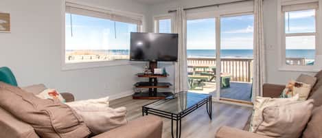 Een woonkamer met een flatscreen televisie, beige banken, een glazen salontafel en uitzicht op het strand door grote ramen.