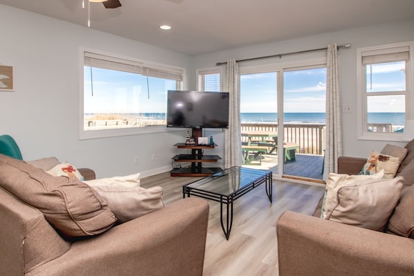 Una sala de estar con televisor de pantalla plana, sofás color beige, una mesa de centro de vidrio y vista a la playa a través de ventanas amplias.