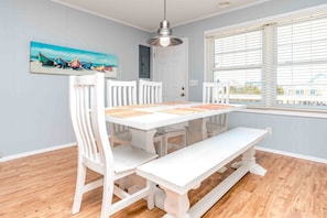 A dining area with a white table and chairs, a painting on the wall, and a window with blinds. - 75 Trips - 3BR 2nd Row Home - Covered Porch & Bonus Space; Dog Friendly! (Topsail Beach)