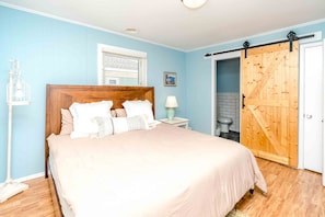 A bedroom with a bed, a nightstand, a lamp, a window, a door, and a view of the bathroom. - 75 Trips - 3BR 2nd Row Home - Covered Porch & Bonus Space; Dog Friendly! (Topsail Beach)
