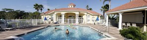 A swimming pool with lounge chairs and umbrellas, surrounded by palm trees and a building with a red-tiled roof.