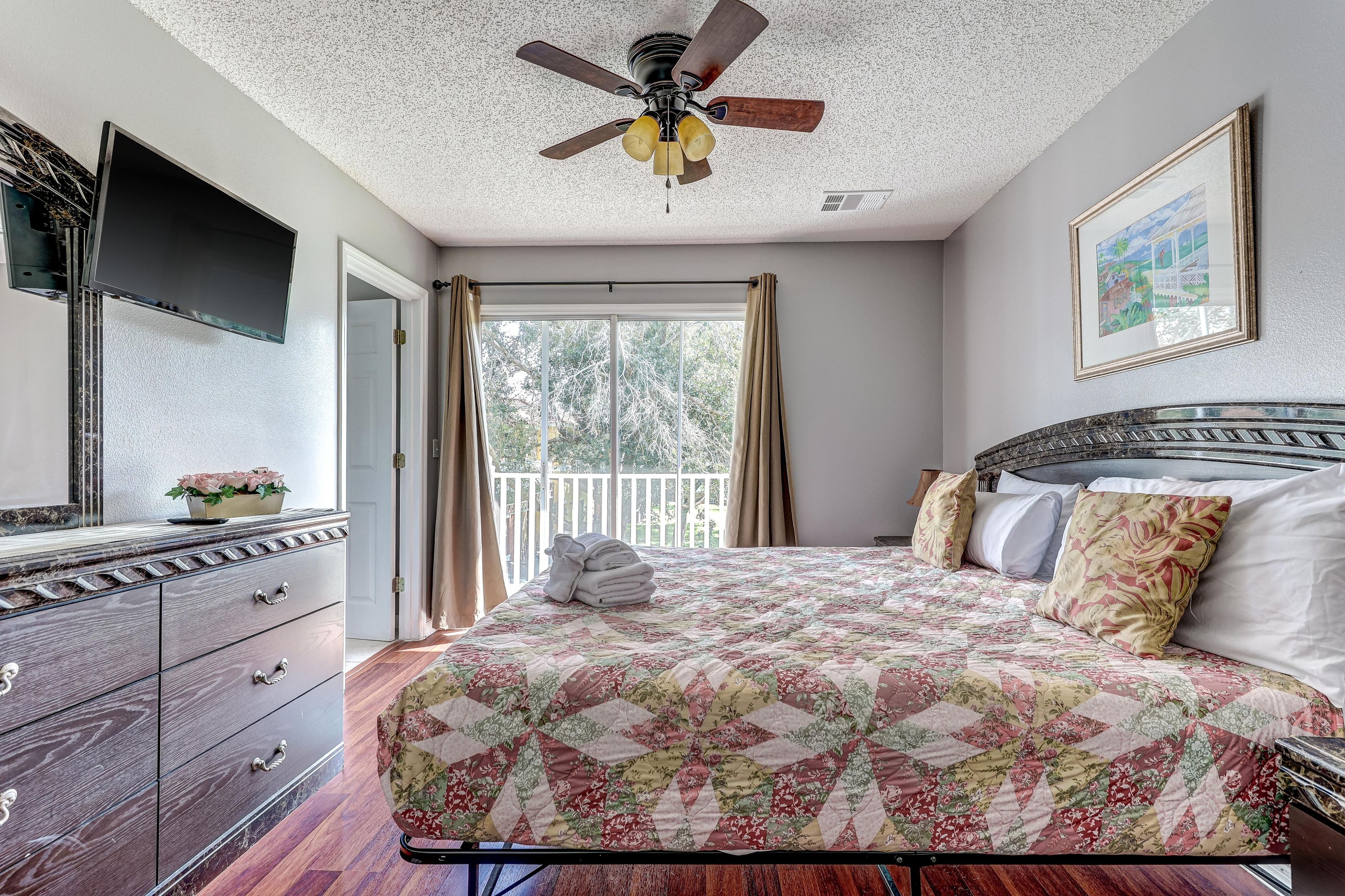 A bedroom with a bed, a dresser, a ceiling fan, a window with curtains, and a painting on the wall.