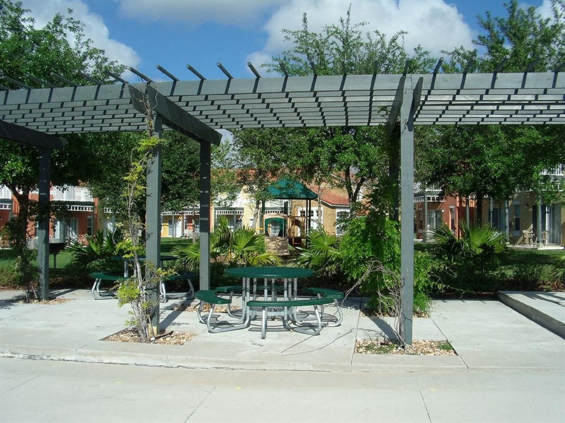 A covered outdoor seating area with tables and chairs, surrounded by greenery and buildings in the background.