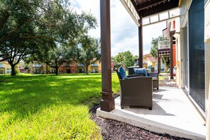 A covered outdoor seating area with wicker furniture, a grassy lawn, and a view of colorful buildings in the background.