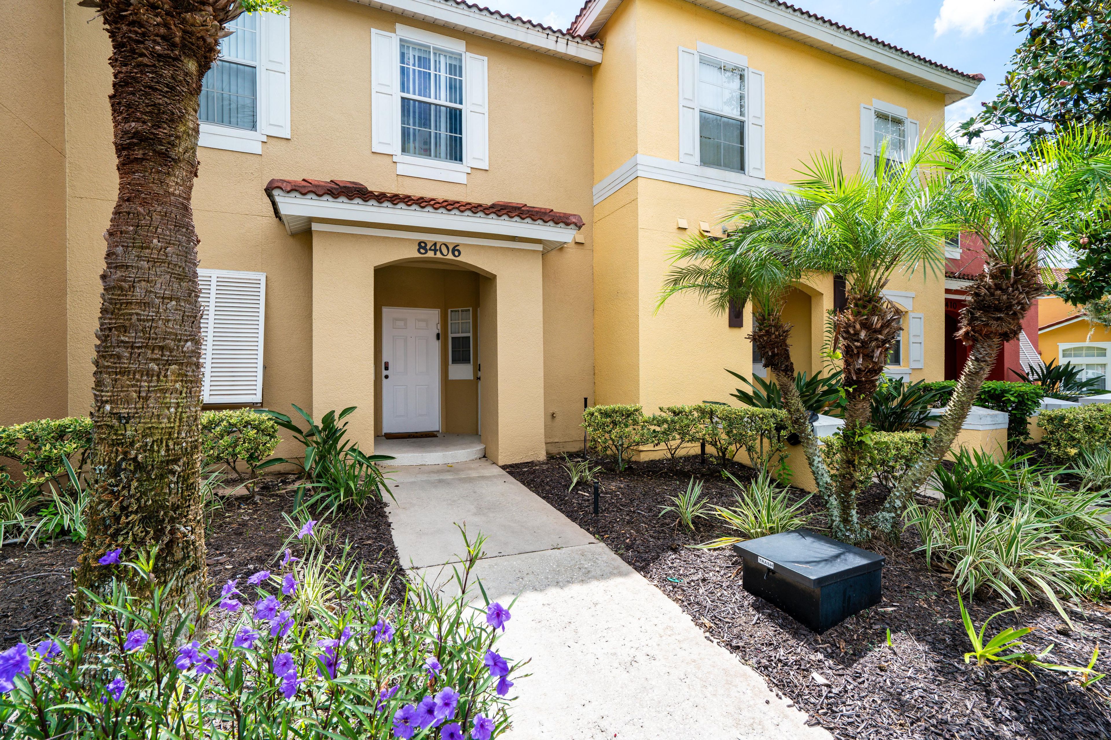 A yellow building with a white door and the number 8406. There are palm trees and purple flowers in the garden.