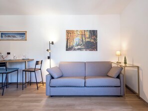 A modern living room with a grey sofa, a dining table with chairs, a painting of a forest, and a floor lamp.