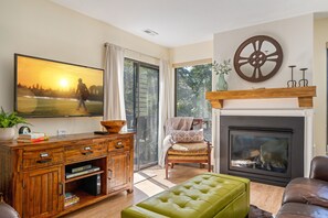 A living room with a fireplace, a large window, a green ottoman, and a wooden cabinet with books. - SKI N SKI OUT. 5 Min Walk to Historic Main St.  Ski, Hike, Bike,  out the door. (Park City)