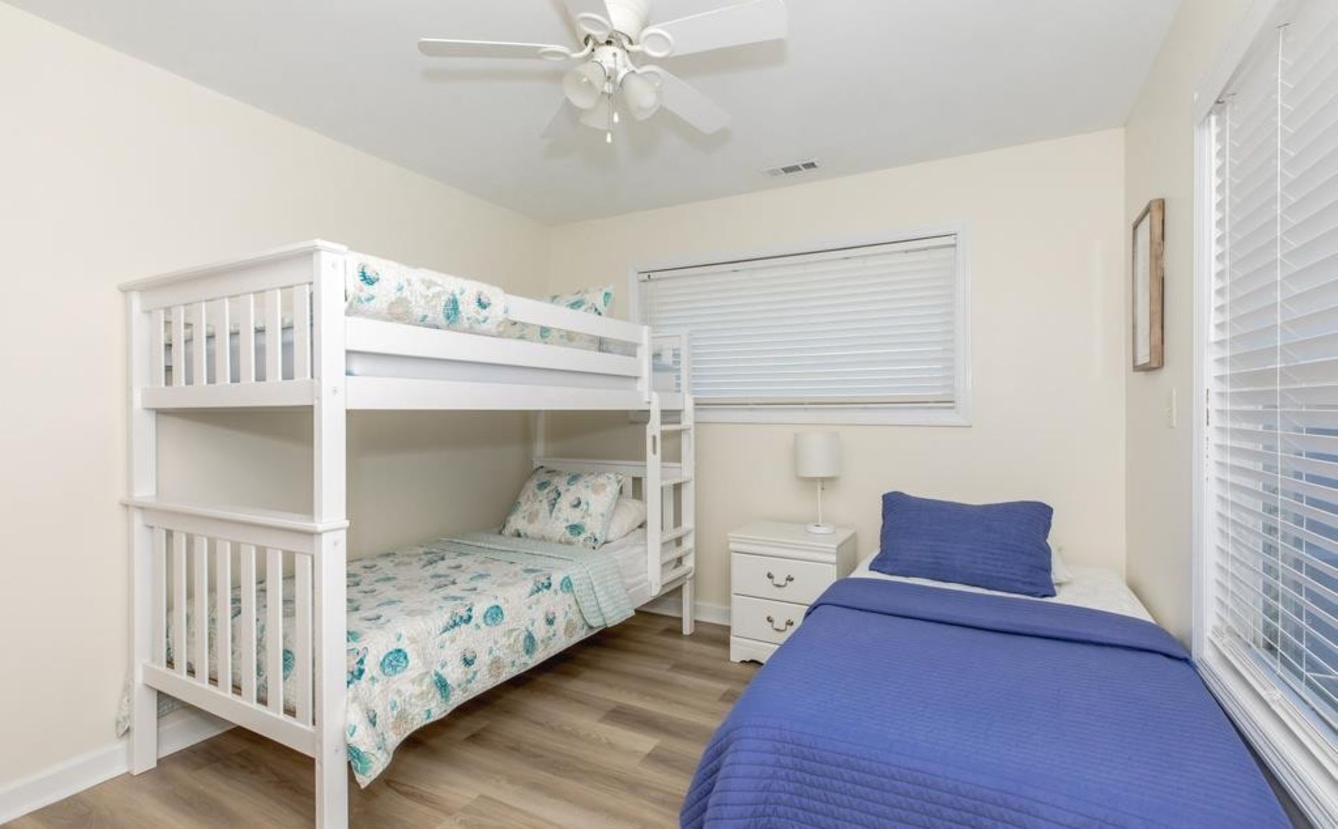 Un dormitorio con una cama litera, una cama individual, una mesita de noche, una lámpara y una ventana con persianas.