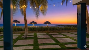 Blick auf einen Strand mit Palmen, strohgedeckten Sonnenschirmen und Lichterketten.