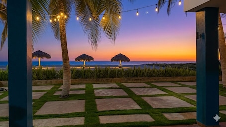 A beach view with palm trees, thatched umbrellas, and string lights.