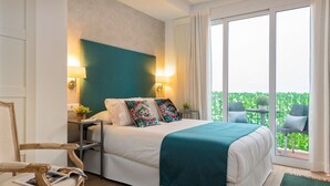 Ein Hotelzimmer mit Bett, Nachttisch, Stuhl, Fenster und einem Balkon mit Grünpflanzen.