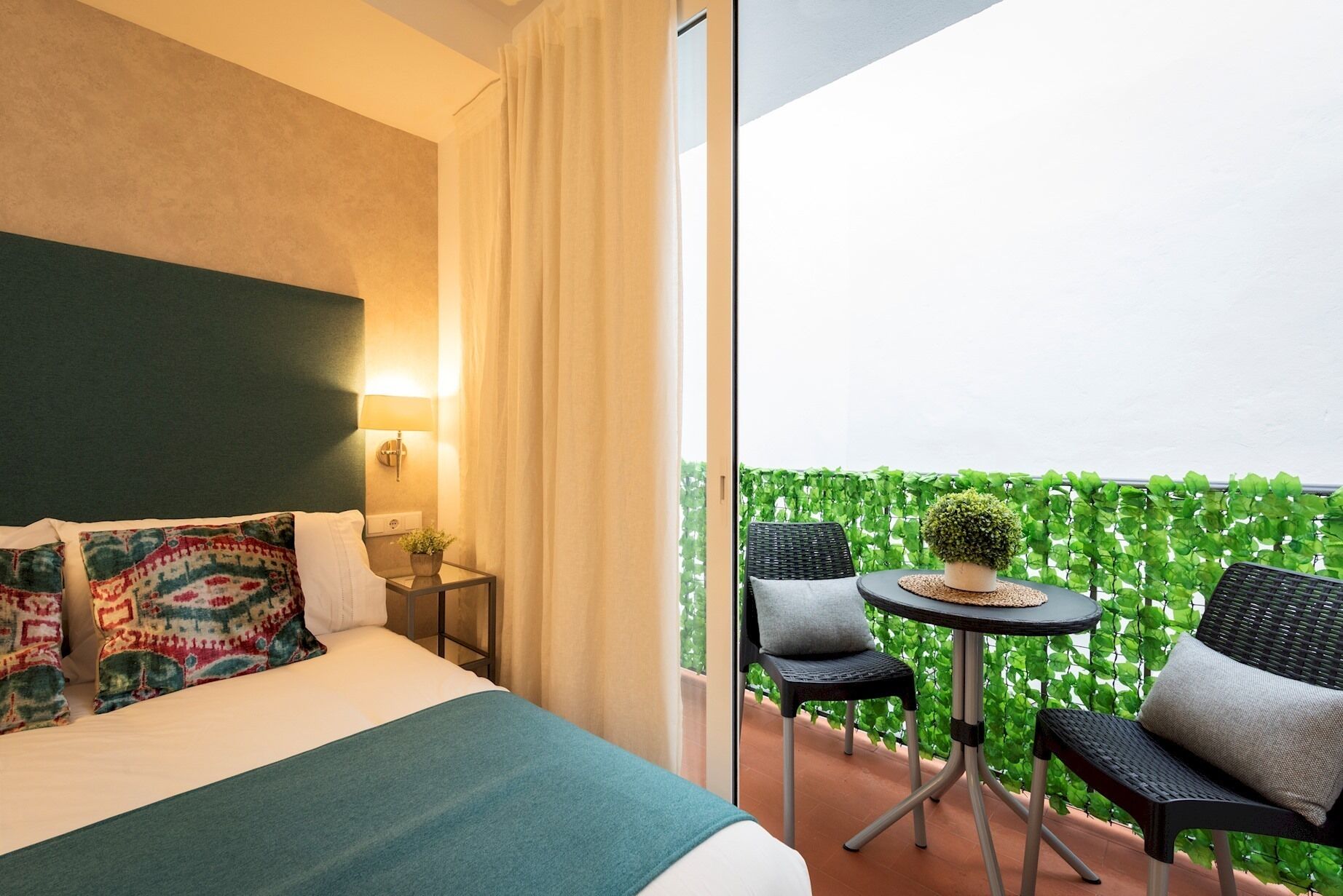 A hotel room with a bed, a small table with chairs, and a view of a green hedge outside.