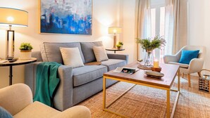 Ein modernes Wohnzimmer mit einem grauen Sofa, einem Couchtisch und einem blauen Sessel. Auf den Nachttischen stehen zwei Lampen, an der Wand hängt ein Gemälde, und ein Fenster ist mit Vorhängen drapiert.