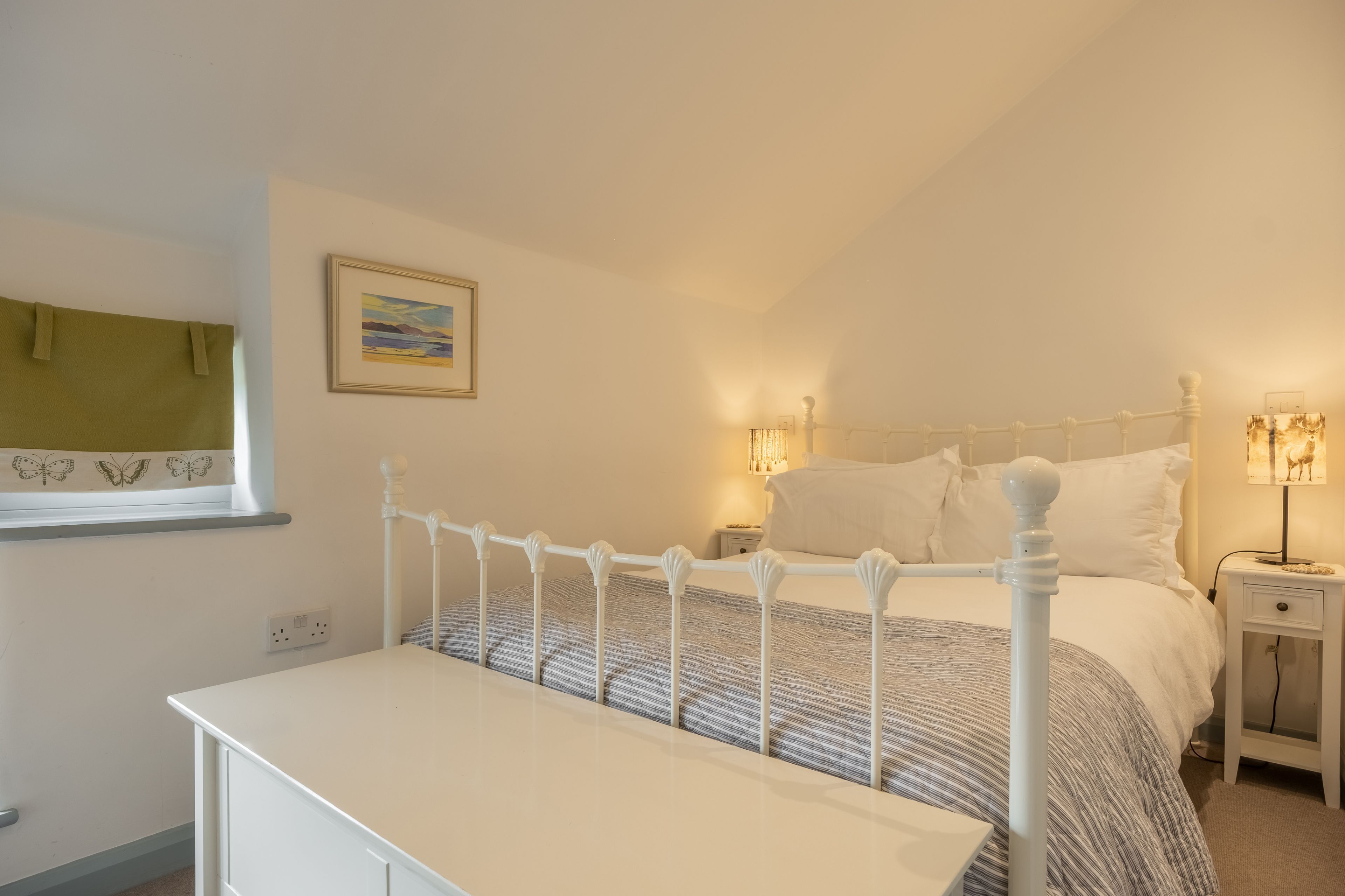 Una camera da letto con un letto bianco, comodini, un tavolo da notte, una finestra con tende e un quadro appeso al muro.