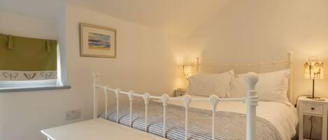 Una camera da letto con un letto bianco, comodini, un tavolo da notte, una finestra con tende e un quadro appeso al muro.